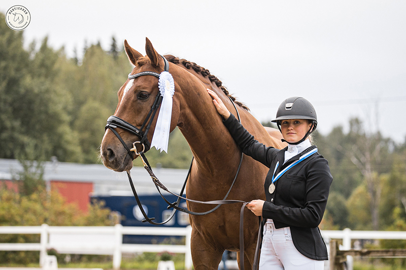 Laura Kangasniemi hymyilee kameralle silittäen kilparatsastusasussa hevosensa kaulaa, omassa kaulassaan mitali ja hevosen suitsissa ruusuke.
